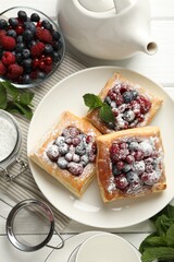 Tasty puff pastries with berries, powdered sugar and mint on white wooden table, flat lay