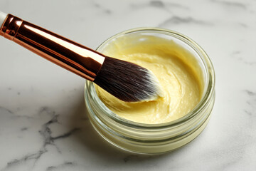 Closeup of a makeup brush dipped in a jar of yellow cream on a marble surface