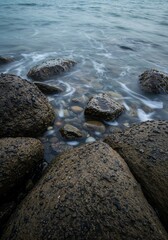 Obraz premium Serene Seaside Stones: A cluster of weathered stones meets the gentle embrace of the tranquil sea, the rhythmic lapping of the waves adding to the peaceful atmosphere.