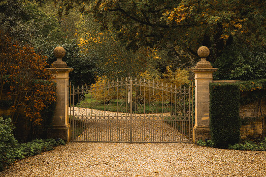 Wrought iron gates closing a gravel driveway leading to a secluded autumn estate. Cotswolds countryside, England