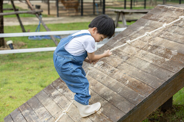 Asian Young boy climbing wooden ramp with rope in outdoor playground, wearing denim overalls and...