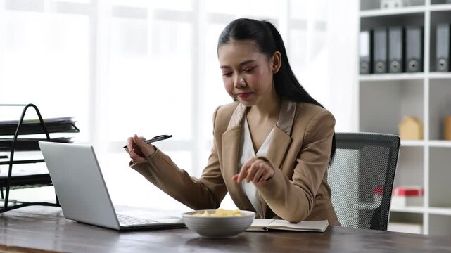 Businesswoman eating potato chips while typing on laptop, blending work and snacking in office with relaxed.
