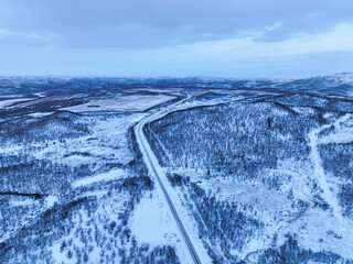 Aerial view of a straight road cutting through the vast, snow-covered forest tundra landscape of the Murmansk region with rolling hills in the distance under a blue twilight sky
