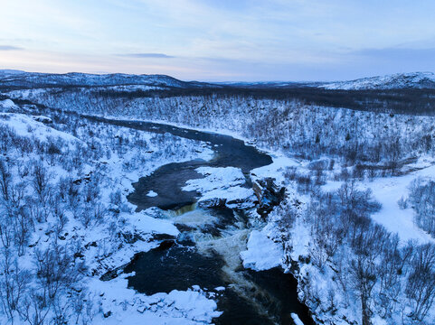Aerial view capturing a cascading river with powerful rapids and a partially frozen waterfall set within a snow-covered forest tundra valley in the Murmansk region