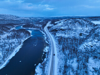 Murmansk, Russia - December 13, 2024: An editorial aerial view of a black Jetour T2 SUV driving on a snow-covered road that runs parallel to a dark, partially frozen river through a cold, mountainous 