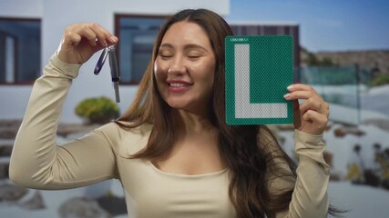 Woman smiling holds car keys and learner sign at house entrance outdoors; pride independence achievement.