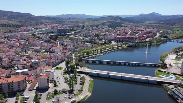 Scenic panorama of Mirandela cityscape on banks of Tua overlooking residential districts, two bridges across river and gentle mountain slopes covered with forests in background on spring day, Portugal