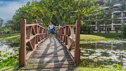 A man stands on a wooden pedestrian bridge over a pond, smiling, holding onto the railing. Leaves, flowers of water lilies on the water surface. Tropical trees, the hotel building in the background. 