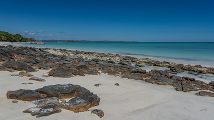 Boulders are scattered on the sandy beach. A calm aquamarine ocean. Motorboats are moored in the distance. Silhouettes of people. Green vegetation, palm trees on the coast. The blue sky. Madagascar.
