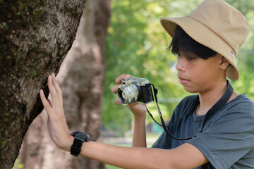 A young photographer in a bucket hat taking a close-up shot of tree bark with a camera, showing...