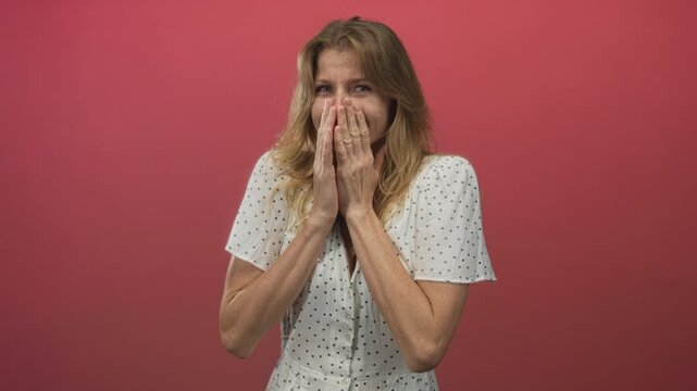 Woman with hands raised and fingers pinched near face in pink studio wearing white polka dot dress; embarrassment.