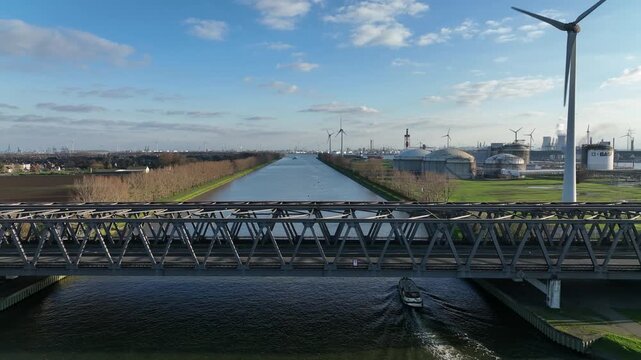 Zoom in aerial shot of a boat crossing under bridge in Antwerp canal surrounded by industrial zone and wind turbines on a sunny day