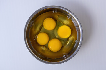 Close-up of four bright yellow egg yolks floating in clear egg white in a steel bowl on a white background. Fresh, protein-rich ingredient for omelette, baking, or frying. Healthy cooking concept.