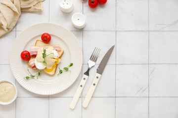 Plate with tasty egg Benedict, tomatoes, bowl of sauce and cutlery on white tile background
