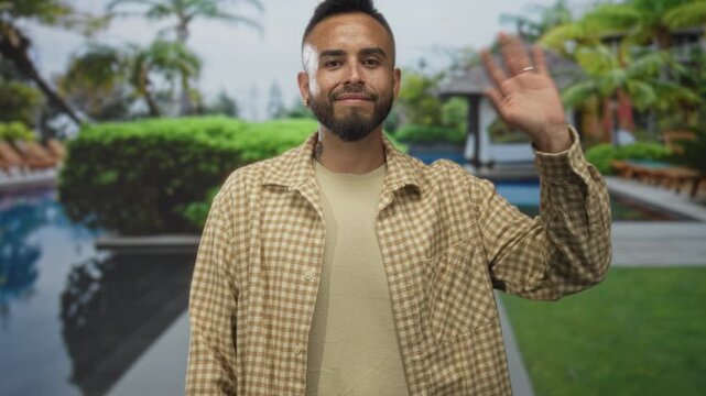 Man waves hand and smiles with face visible at resort, wearing checked shirt and ring on hand; vacation serenity.