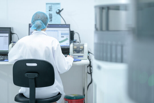 From behind, a lab professional in a hair net works at a computer, analyzing patient test results and monitoring medical equipment in a modern hospital laboratory. - Powered by Adobe