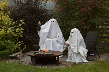 mom and kid in Halloween ghost costumes roast marshmallows on the fire near the house. Halloween party