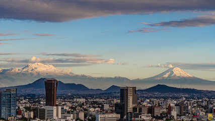 A city with mountains in the background. The sky is blue and the clouds are white. Panoramic view of Mexico City, snow-covered volcanoes Popocatepetl and Iztaccihuatl