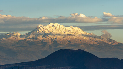 A mountain range with snow on the top and a cloudy sky. The mountains are in the distance and the sky is blue. Panoramic view of Mexico City, snow-covered volcanoes Popocatepetl and Iztaccihuatl