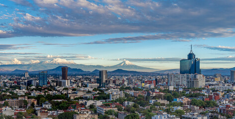A city with a mountain range in the background. The sky is blue and there are some clouds. Panoramic view of Mexico City, snow-covered volcanoes Popocatepetl and Iztaccihuatl © Alejandro