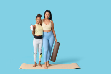 African-American mother with her teenage daughter holding yoga mat and water bottle on blue background