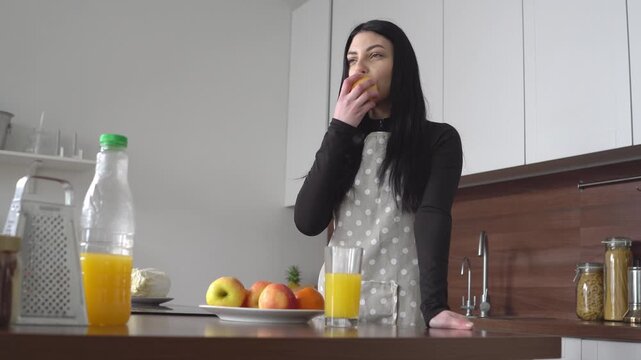 young woman in apron eats apple in the kitchen, girl bites the fruit and relaxes at home
