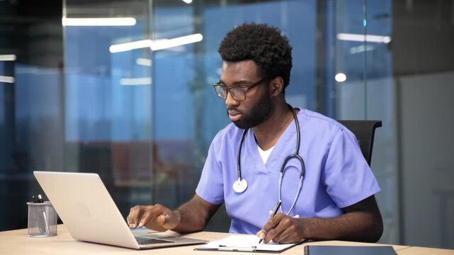 Busy african american doctor in blue coat working at workplace taking notes using computer in hospital office. Medical worker physician doing paperwork, filling out form or writing out prescription
