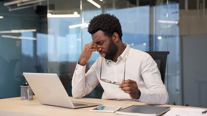 Tired african american businessman suffering from eye strain while working on a computer sitting at workplace in business office. Overworked male takes off glasses and massages, rubs bridge of nose