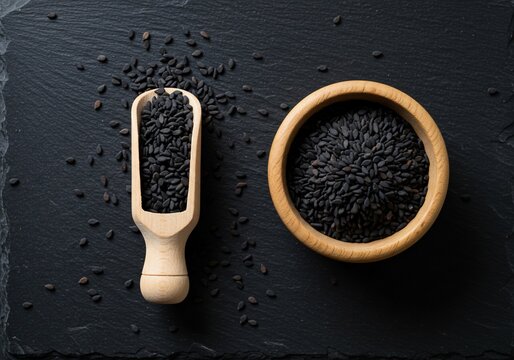 Black cumin seeds in a wooden bowl and scoop on a dark slate background top view.