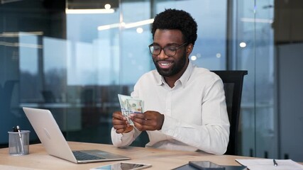Happy african american businessman enjoys successful financial gain, counting cash with satisfaction while sitting at desk at workplace in business office. Male worker calculates his earnings, salary - Powered by Adobe