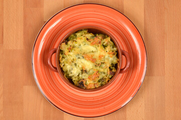 A portion of baked Brussels sprouts and potato with melted cheese in a small ceramic cocotte, served on a rustic orange plate on a wooden surface