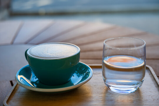 A teal ceramic cup of frothy coffee with a glass of water rests elegantly on a wooden tray atop a patterned caf&eacute; table.