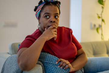 Worried stressed african american woman sitting on sofa at home, deep in negative thought. Pensive frustrated black female with hand on chin reflecting on life problems, feeling anxious and uncertain.