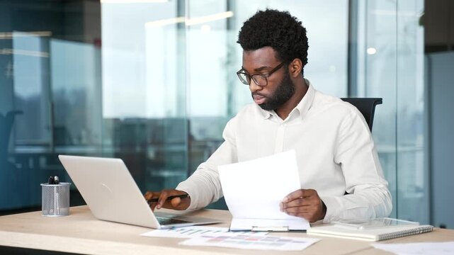 Busy african american businessman is doing paperwork using computer while sitting at workplace in business office. Thoughtful financier working with documents, making a report, engaged in accounting