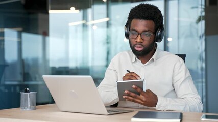 African american businessman in headphones watching video call conference takes notes looking at laptop screen sitting at workplace in business office. Focused worker listening online training, course