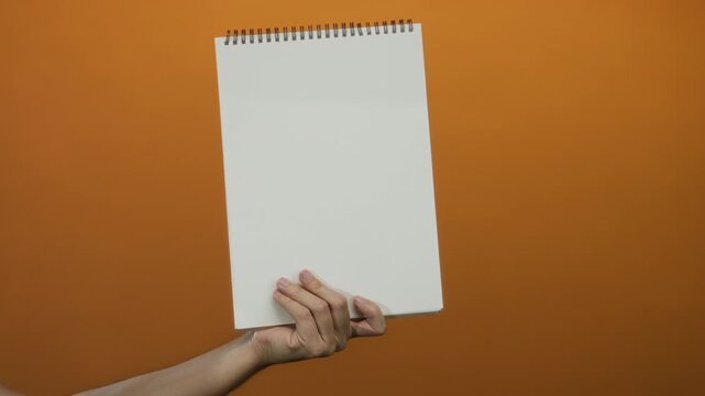 Man holding blank notepad with white pages against orange wall background, emphasizing creative space and isolated ideas in a minimalist setting.
