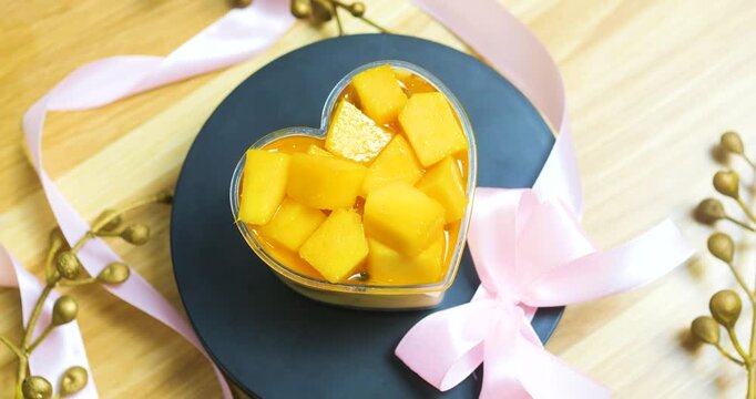 Heart shaped mango dessert sprinkled with golden accents, tied with pink ribbon, resting on dark plate against wooden background, symbolizing romantic sweetness
