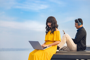 Time to relax. Handsome middle-aged asian man in wireless headphones watching videos on mobile phone. Asian woman busy using laptop surfing wireless web shopping online while relaxing at beach. 