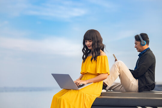 Time to relax. Handsome middle-aged asian man in wireless headphones watching videos on mobile phone. Asian woman busy using laptop surfing wireless web shopping online while relaxing at beach. - Powered by Adobe