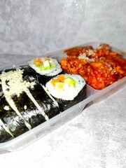 A plastic take-out container holding a Korean combo meal with sliced kimbap rolls topped with mayonnaise and grated cheese, alongside a portion of spicy red-sauced chicken (Dakgalbi or Yangnyeom).