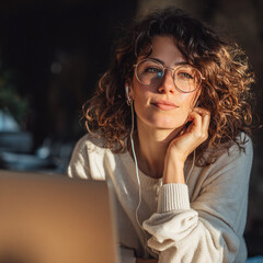 Young woman with curly hair and glasses smiling softly while working on laptop – warm golden hour lighting, cozy home office vibe, authentic remote work concept 