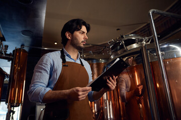 A man wearing an apron is studying a clipboard in a brewery setting