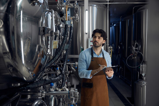 Brewer wearing an apron is holding a clipboard inside a brewery facility