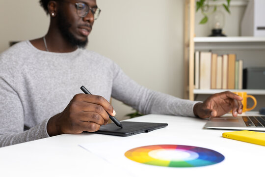 Close up of African American male graphic designer working at home using laptop and graphic tablet. Selective focus.