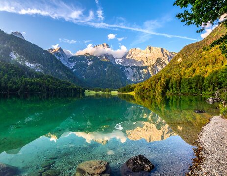 Scenic mountain lake reflects forest and snow-capped peaks under a bright, partly cloudy sky