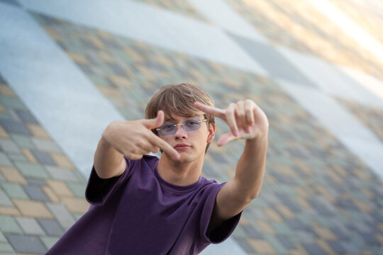 Male youth in purple shirt poses with hands framing his face, displaying a confident and playful demeanor against a backdrop of geometric patterns and textures