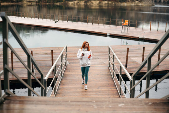 Sporty woman holding smartphone walking wooden pier near lake