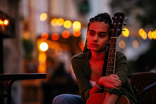 teenager with braids plays an acoustic guitar on an evening street.