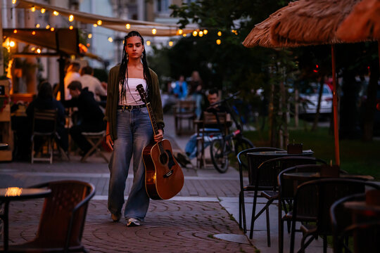teenager with braids with guitar walking on city street at evening.