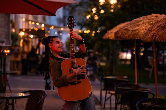 teenager with braids plays an acoustic guitar on an evening street.